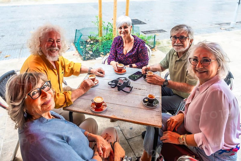 Smiling group of senior friends gathered around an outdoor cafe table, enjoying coffee, conversation and relaxed togetherness on a sunny city day, diverse and joyful