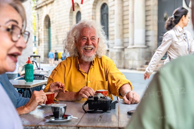 Mature man with white hair and beard laughing brightly while enjoying coffee and conversation with friends at an outdoor cafe, sharing moments of joy and connection in an urban setting