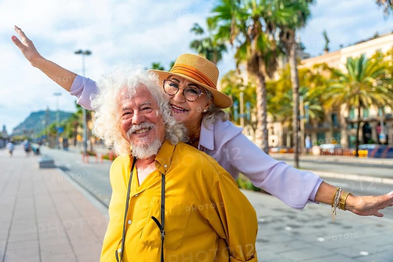 Senior couple laughing as husband gives wife a piggyback through sunlit city streets lined with palm trees, enjoying a carefree vacation together and making joyful memories