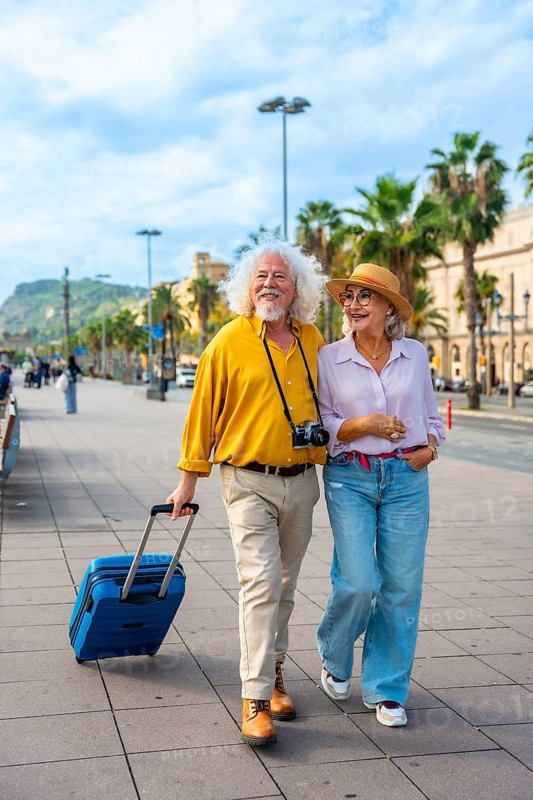 Smiling senior couple embracing a happy retirement, walking together with their luggage through a sunny city street, exploring and enjoying an exciting travel adventure