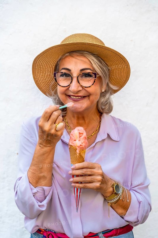 Happy senior woman wearing a straw hat and glasses smiling while eating a refreshing ice cream cone with a small spoon, celebrating moments of joy and relaxation in summer
