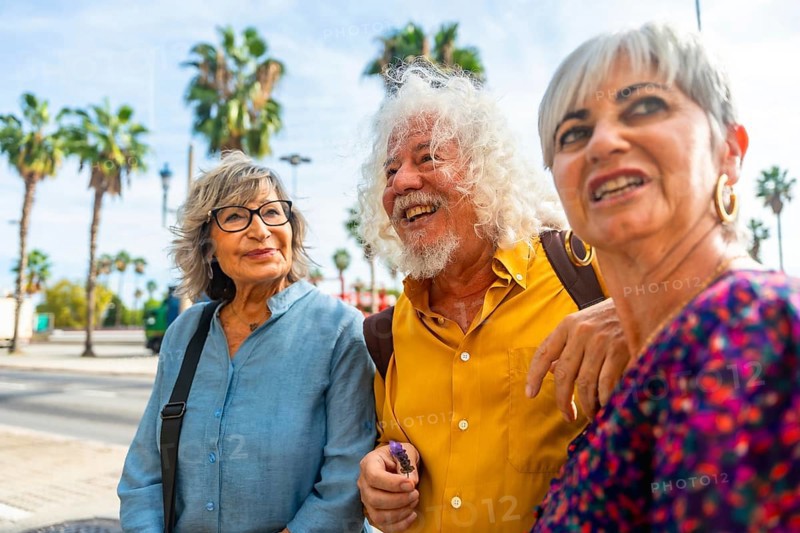 Three happy senior friends enjoying a vacation while walking together on a city street, sharing moments and discussing, with palm trees visible in the background under a clear sky