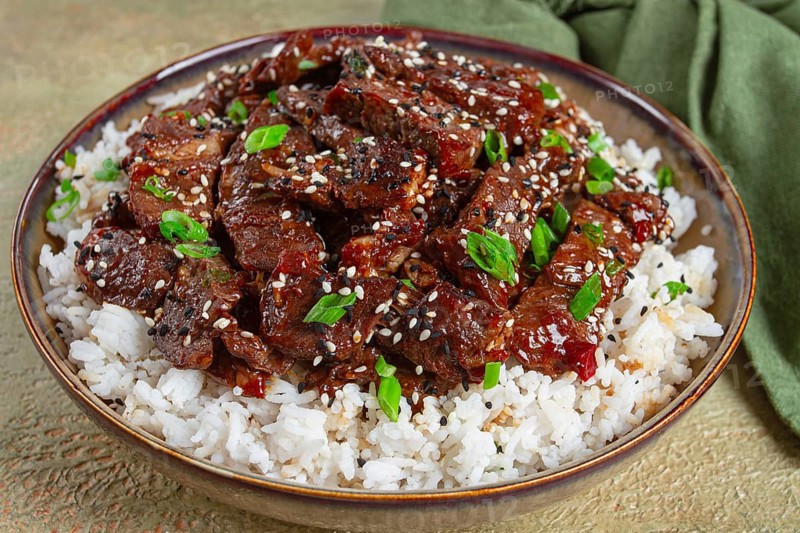 Crispy beef, with green onions and sesame seeds, on a rice pad, in a bowl with chopsticks, on a textured surface, no people