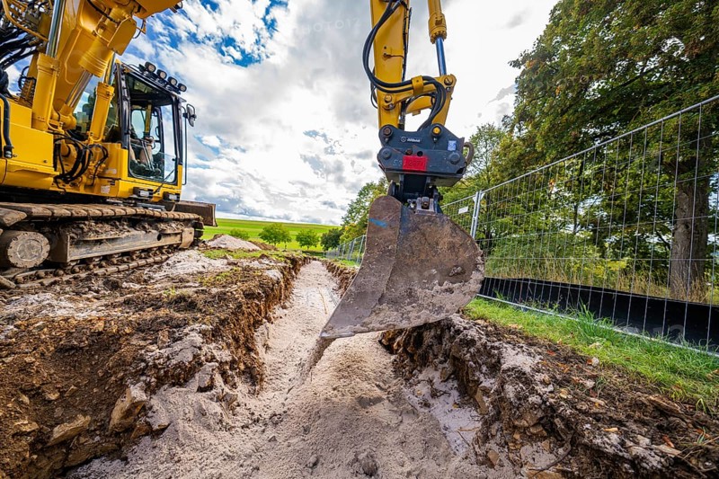 An excavator lifts soil from a deep ditch on a construction site in cloudy skies, construction PV Freifaechenanlage, Weil der Stadt, Germany