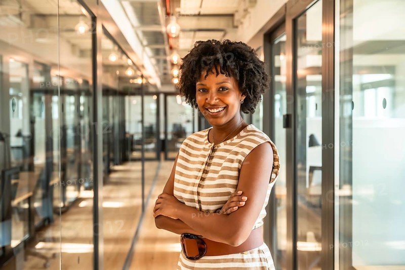 Black businesswoman standing with folded arms and a confident smile, working in a contemporary coworking space hallway with glass walls and warm lighting