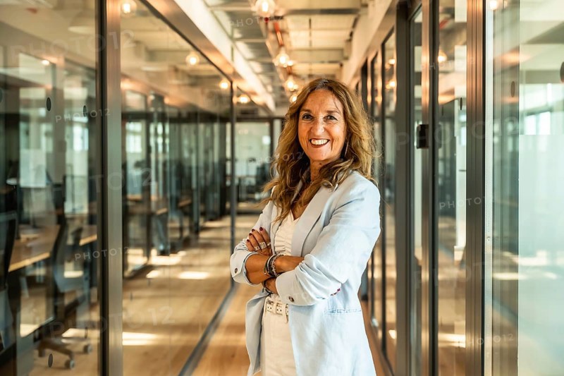 Mature businesswoman standing with folded arms in a bright modern office corridor, exuding confidence, professionalism, and leadership in the workplace
