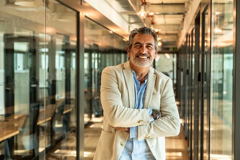 Mature executive standing confidently with arms crossed in a contemporary office corridor, celebrating success and projecting a professional and approachables