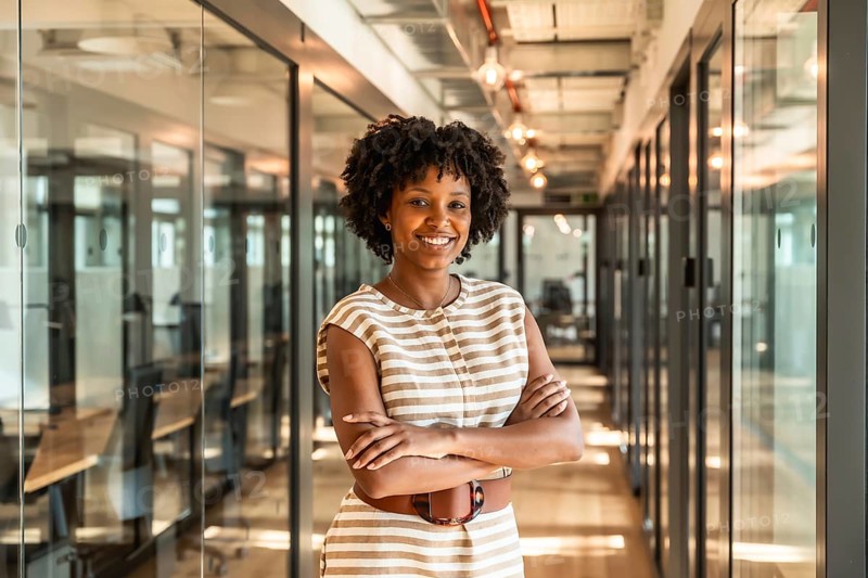 Young african american businesswoman smiling with crossed arms, standing confidently in a modern coworking office corridor with glass walls and bright lighting