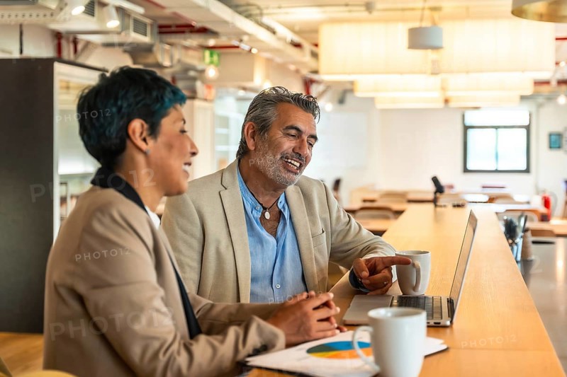 Diverse business colleagues are collaborating on a laptop, sharing ideas and discussing data in a bright coworking space, fostering teamwork and professional interaction