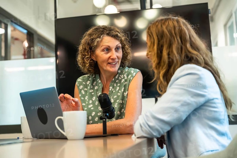 Two professional women collaborating on a podcast, recording a conversation with a microphone and laptop in a contemporary coworking space, focusing on content creation and communication