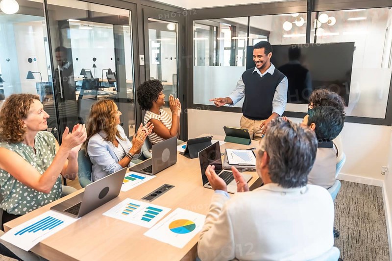 Diverse business team in a modern boardroom applauding a smiling presenter, celebrating project success and teamwork during a collaborative meeting with laptops and charts on the table