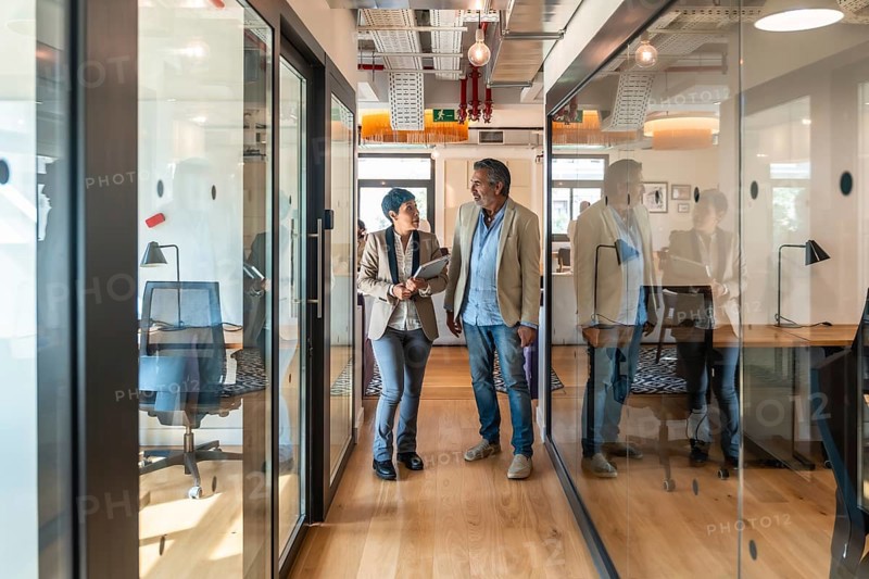 Two diverse business colleagues, a man and a woman, walking along a glass walled corridor in a contemporary office while discussing work and holding a tablet