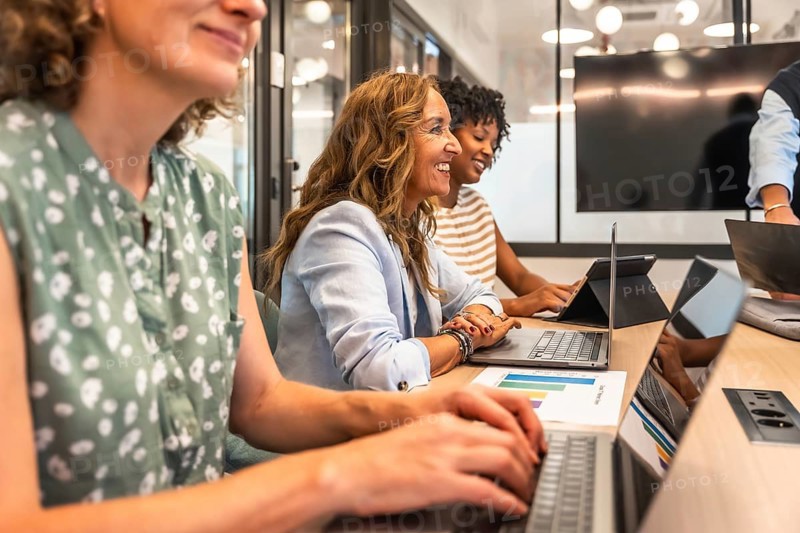Diverse group of smiling businesswomen collaborating and engaging in productive work during a team meeting, using laptops and reviewing data in a contemporary coworking office environment