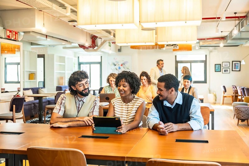 Diverse business colleagues collaborating around a tablet in a modern open plan office, brainstorming ideas, sharing plans and smiling during a productive daytime meeting