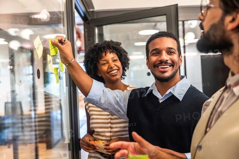 Diverse business professionals collaborating in a modern office, cheerfully sharing innovative ideas during a meeting, placing colorful sticky notes on a glass board