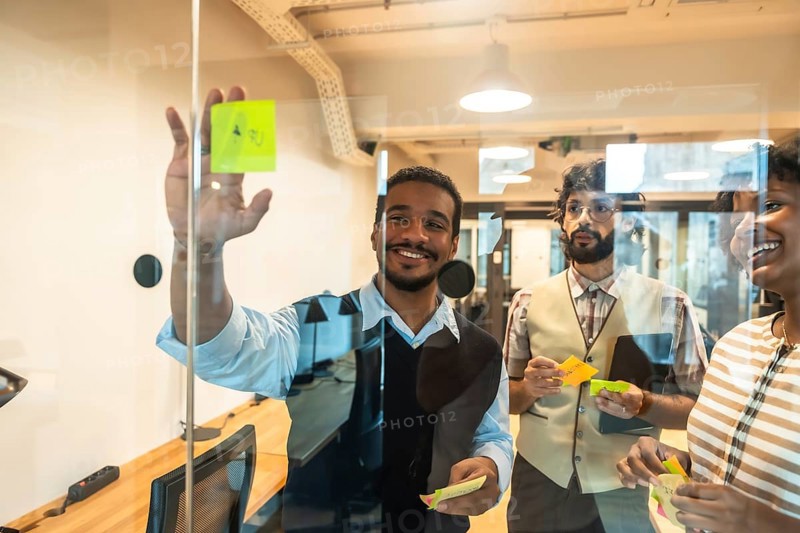 Happy diverse business team collaborating on glass wall during brainstorming session, sharing ideas and strategizing using colorful sticky notes in a contemporary workspace