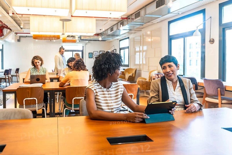 Diverse colleagues engaged in conversation and working on digital devices while collaborating at a shared table in a contemporary, open plan office environment during a business meeting