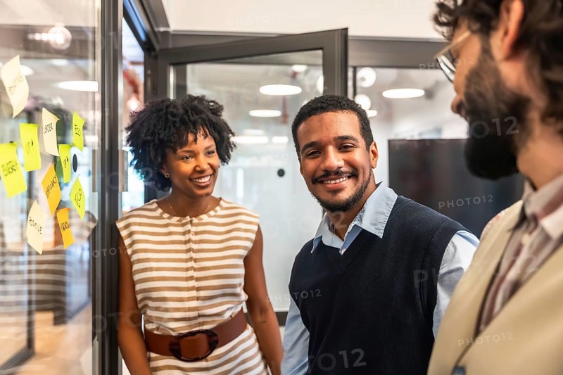 Coworkers smiling and engaging in discussion, brainstorming ideas on a glass board with sticky notes, representing teamwork, diversity, and successful business collaboration