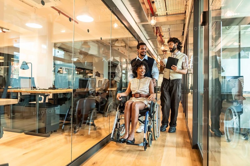 Diverse business colleagues pushing a young black woman in a wheelchair through a modern, accessible office hallway, fostering an inclusive and supportive work environment