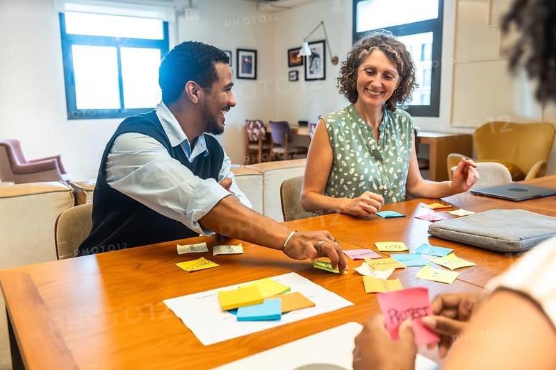 Diverse coworkers sitting around a wooden table, smiling and collaborating on new project ideas using colorful sticky notes during a brainstorming session in a modern office