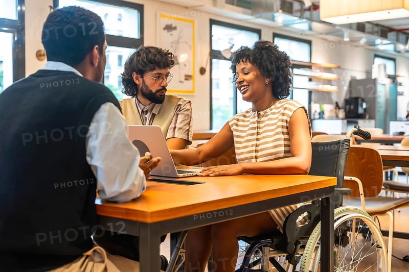 Diverse colleagues collaborating around a modern coworking table, smiling and brainstorming with a laptop, inclusive team includes a woman in a wheelchair, promoting accessibility and teamwork