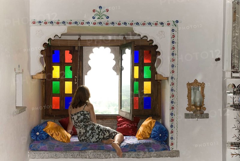 Woman wearing a dress in a cozy sitting area with pillows in front of colorful window, Palasthotel Karni Fort Bambora, Bambora, Rajasthan, India