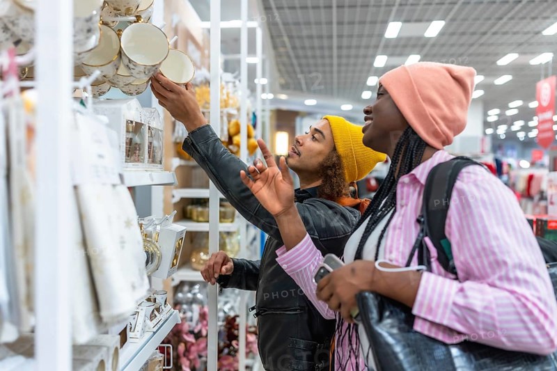 Mixed-race couple browsing colorful home decor and dinnerware on bright store shelves, smiling and enjoying a shared shopping experience while selecting mugs and kitchenware
