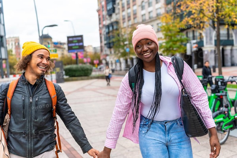 Young multiracial couple smiling and holding hands while strolling down a city street lined with autumn trees, enjoying a casual, cozy outdoor date and relaxed urban togetherness