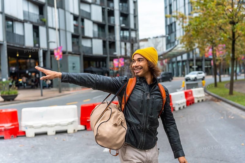 Happy young multiracial man wearing a yellow beanie and leather jacket pointing with his hand while walking and carrying a backpack on an urban street with modern buildings