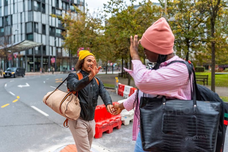 Young diverse couple extending hands for a high five, holding hands, and carrying bags while walking on a city street, representing friendship, travel, and urban exploration