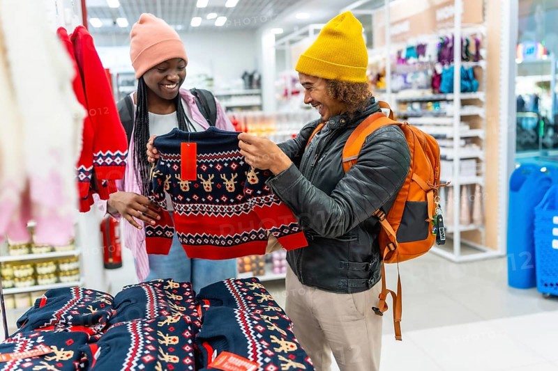 Young couple browsing festive reindeer sweater in a bright store, smiling as they choose holiday clothing together during joyful seasonal shopping and gift hunting
