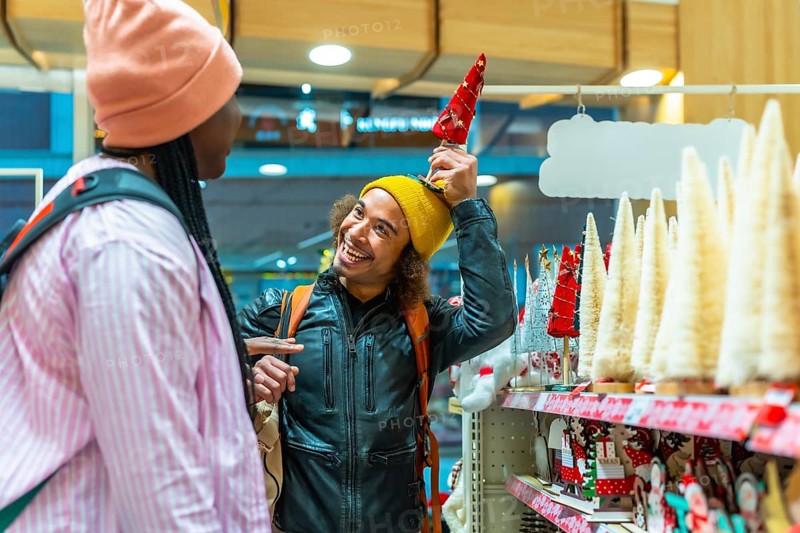 Diverse young couple laughing and shopping for festive holiday decorations in a bright store, enjoying playful togetherness and joyful winter season gift buying and merriment