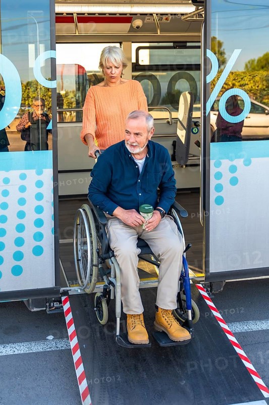 Senior woman is confidently assisting a mature man in a wheelchair to exit a public bus using the extended ramp, demonstrating care and accessibility in urban transportation