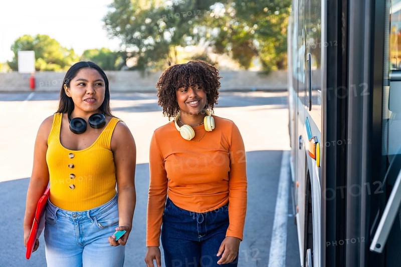 Two multiracial young women standing at a city bus door on a sunny day, smiling and chatting as one holds a folder and headphones, commuting or heading to campus together
