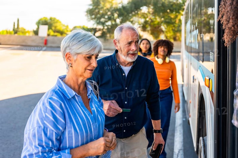 Senior couple approaching a bus entrance, showing a man holding a ticket while waiting in line with other diverse passengers for public transportation