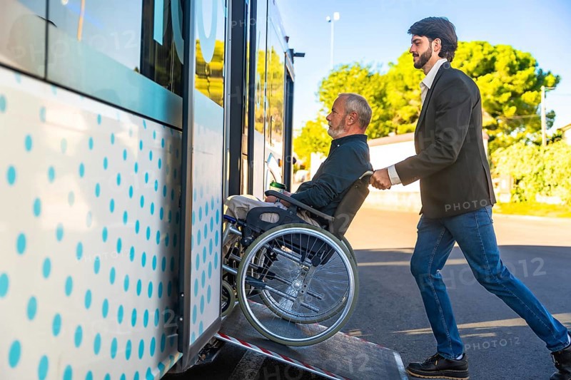 Young man assisting an elderly person using a wheelchair to board a public transport bus via an accessibility ramp, highlighting concepts of care, mobility, and community support