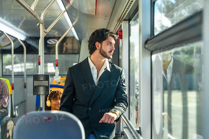 Young businessman in a suit jacket stands by a transit window, looking out pensively as the city moves by during his daytime commute on public transportation