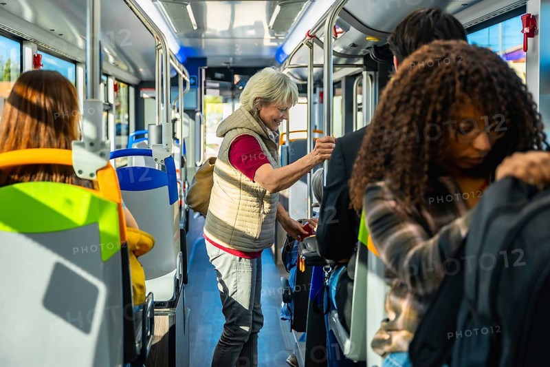 Senior woman standing inside a crowded city bus, holding a handrail among diverse commuters during rush hour urban transit, daily commute and public transportation scene