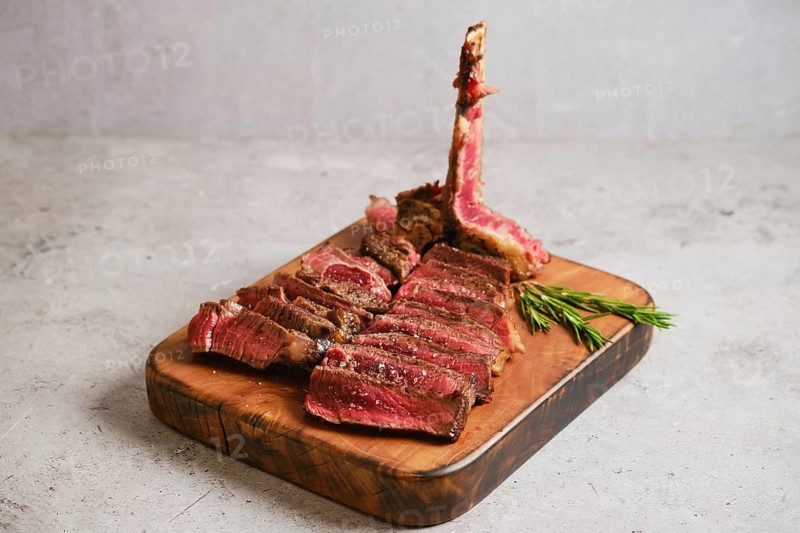 Juicy t-bone beef steak is neatly sliced and arranged on a wooden serving board. The rich red color of the meat contrasts with the light background, showcasing the texture and quality of the beef