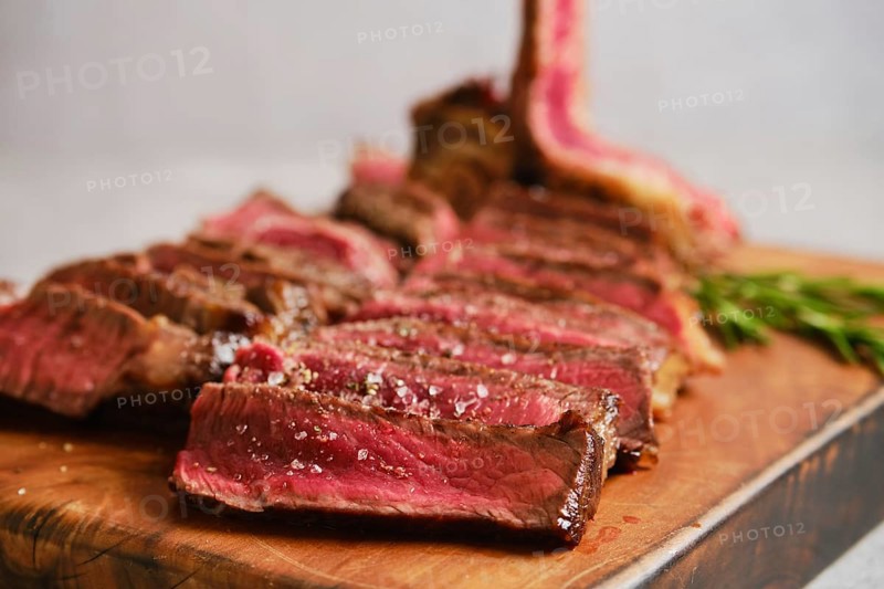 Close-up view of sliced grilled t-bone steak medium rare on a wooden cutting board. The meat is perfectly cooked with a juicy pink center. Fresh herbs and seasoning add color and flavor to the presentation