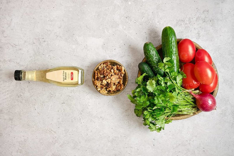 A colorful arrangement of fresh tomatoes, cucumbers, and herbs on a light gray surface. A bowl of crunchy walnuts and a bottle of wine vinegar complete the group, ideal for salad making