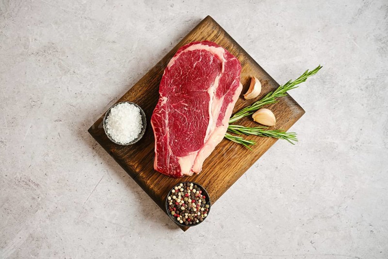 Raw ribeye roll steak rests on a wooden cutting board alongside garlic cloves and fresh rosemary. Nearby, bowls contain sea salt and a mix of colorful peppercorns, ideal for seasoning