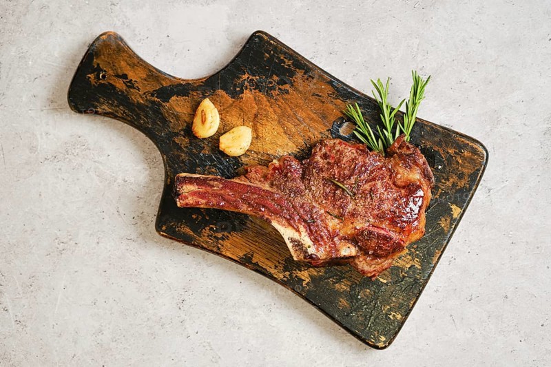 Grilled cowboy steak on a worn wooden cutting board, garnished with fresh rosemary and garlic. The rich brown color of the meat highlights its delicious sear, inviting a tasty dining experience