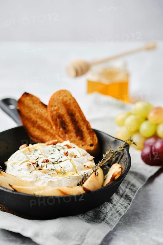 A baked cheese dish is elegantly presented in a cast iron skillet alongside toasted bread and fresh fruits. Honey in a jar adds a sweet touch to this appetizing setup, perfect for a cozy gathering