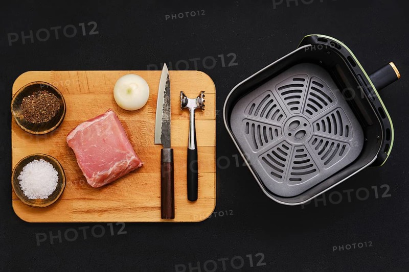 Raw pork tenderloin on a wooden cutting board alongside seasonings and kitchen tools. An onion, salt, and pepper are ready for flavoring before cooking in an air fryer