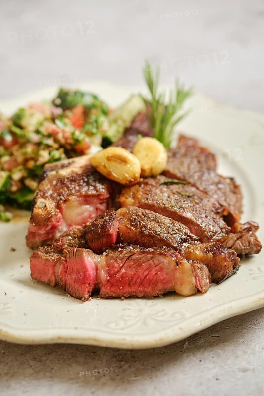 Juicy grilled steak rests on a decorative plate, served alongside a fresh salad made with vibrant greens, tomatoes, and grains. The dish is garnished with garlic for added flavor