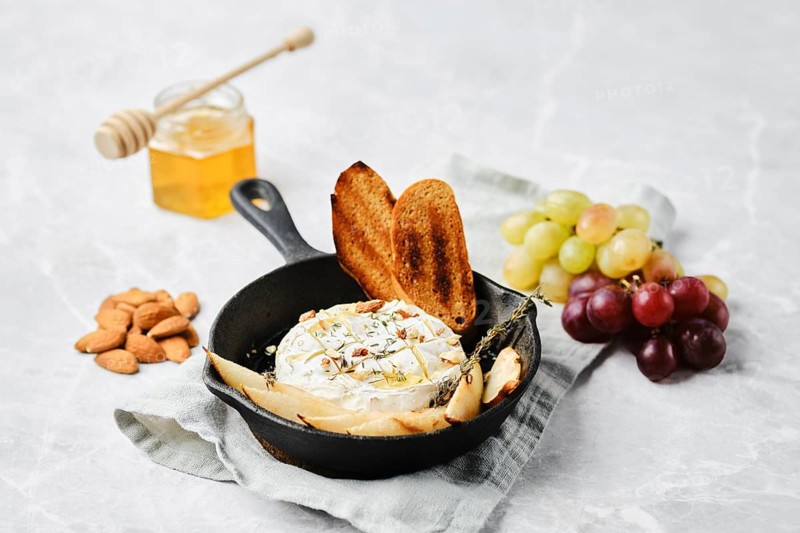 A cast iron skillet holds warm baked cheese, garnished with herbs. Nearby are toasted bread slices, almonds, grapes, and a honey jar, creating an inviting spread for sharing and enjoyment