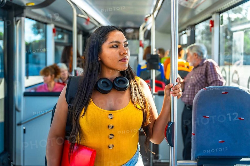 Young woman passenger holding a handrail inside a public bus, wearing headphones and a backpack, looking away while traveling, representing urban daily commute and student life