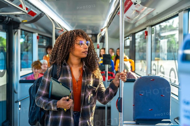 Young woman with curly hair and glasses stands inside a city bus holding a pole, wearing a backpack while commuting through urban streets during a daytime journey