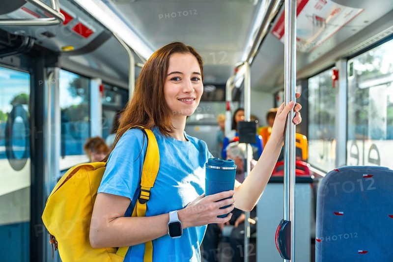 Young woman smiling on a city bus, standing and holding a pole with a yellow backpack and reusable cup, eco conscious commuter enjoying a daytime urban journey